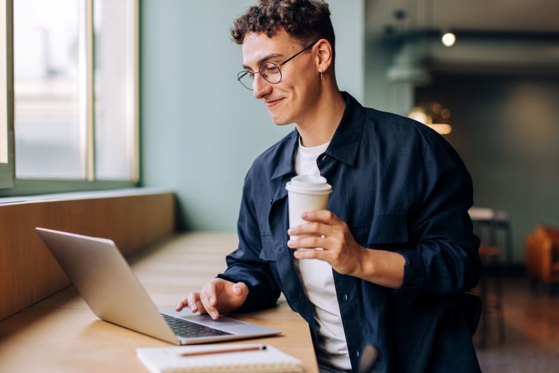 young professional man with brown curly hair working on his laptop and holding a cup of coffee
