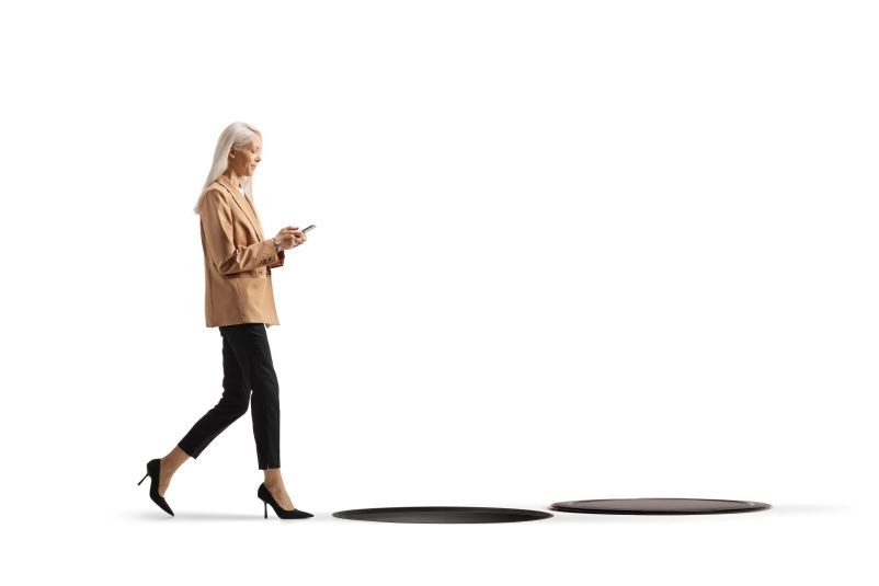young professional woman against a white background, looking at her phone and nearly stepping into a pit in the ground
